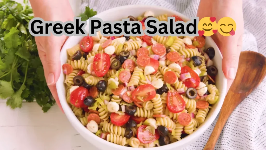 A bowl of Greek pasta salad with pasta, tomatoes, cucumbers, olives, and feta, drizzled with olive oil and herbs.